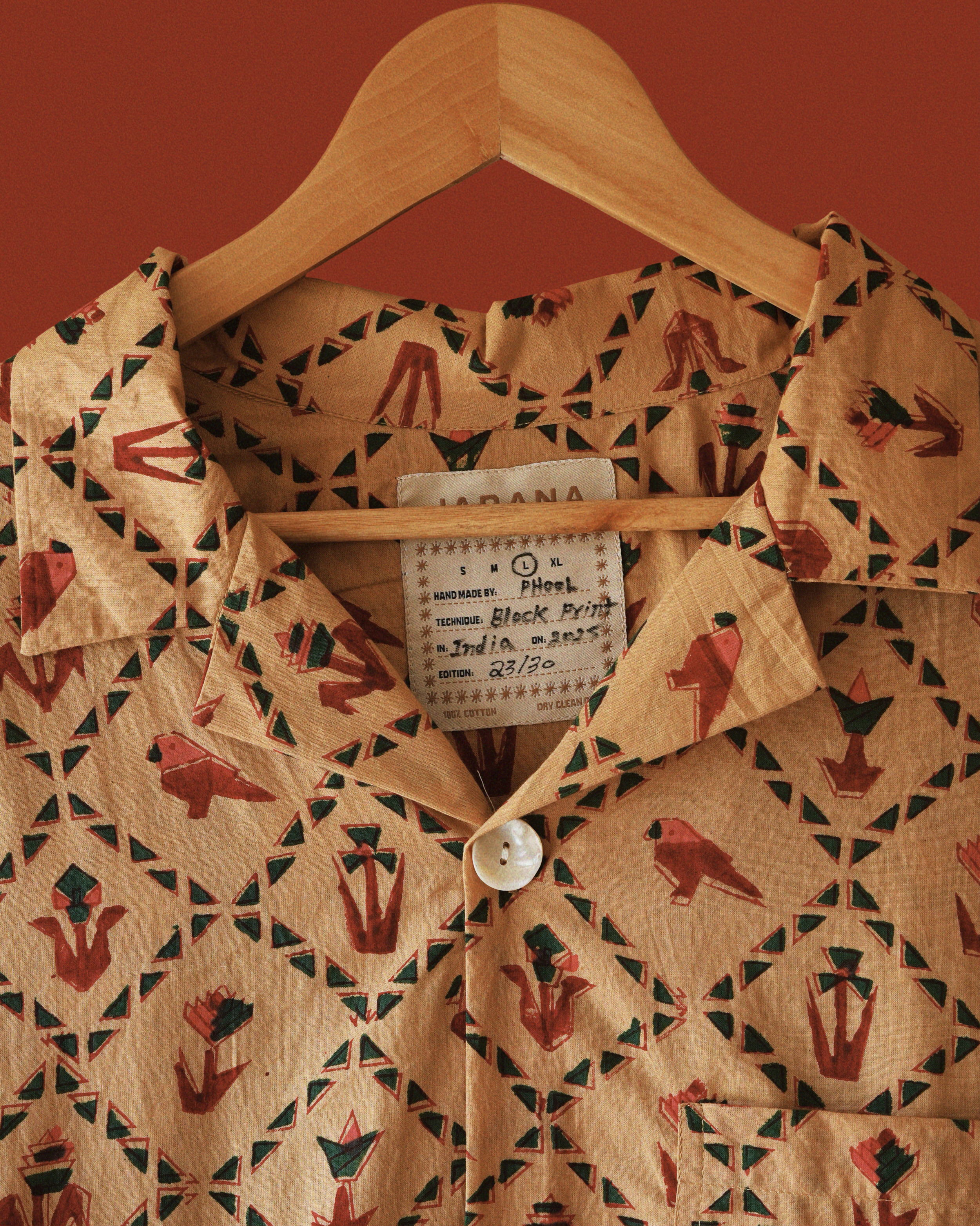 Detailed view of Carioca shirtβs resort collar and Jarana label, showing red parrot and green diamond block-print motifs on sandy cotton.