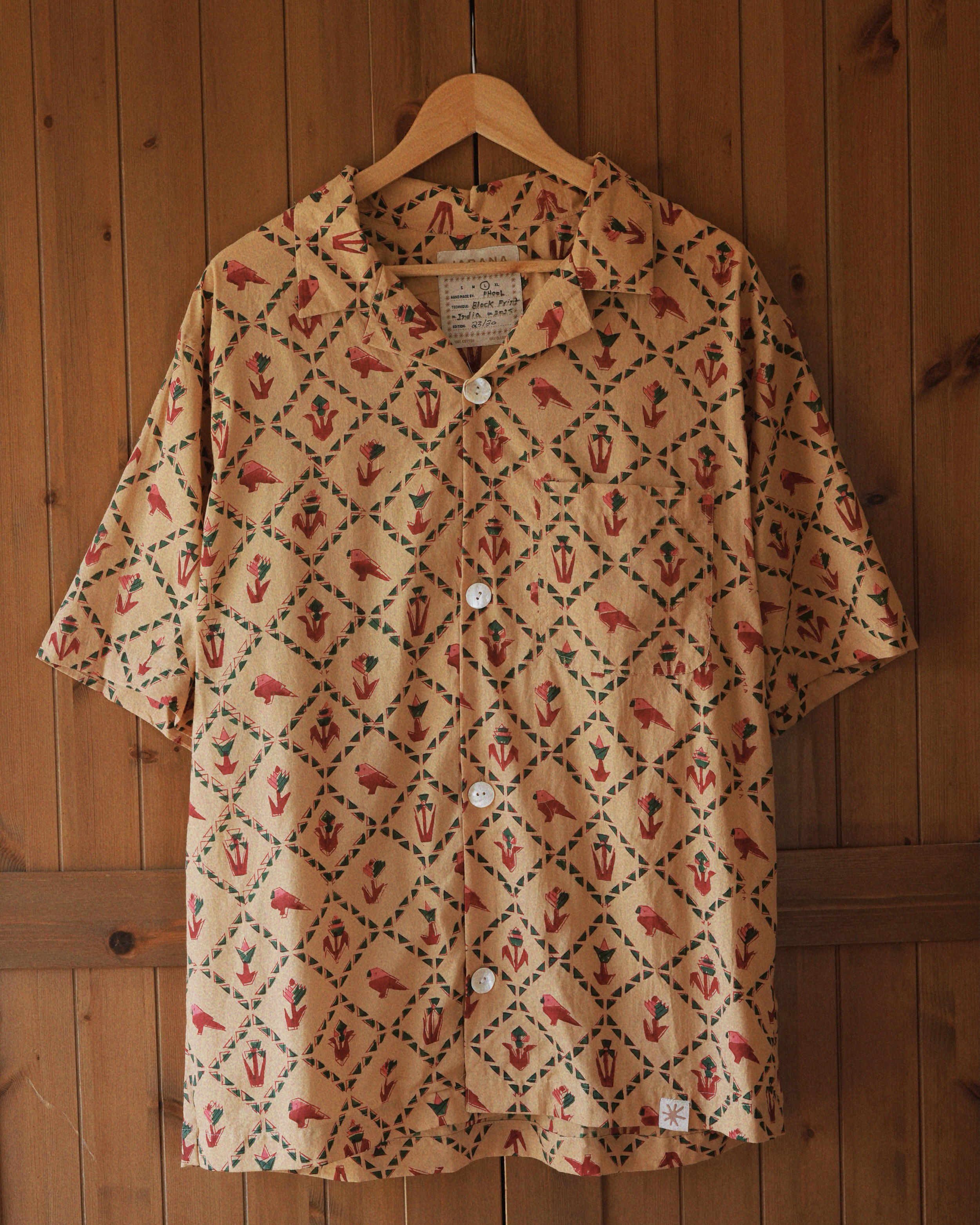 Full front view of Carioca short-sleeve cotton shirt featuring all-over red parrot and green geometric block print, hanging on wooden door.