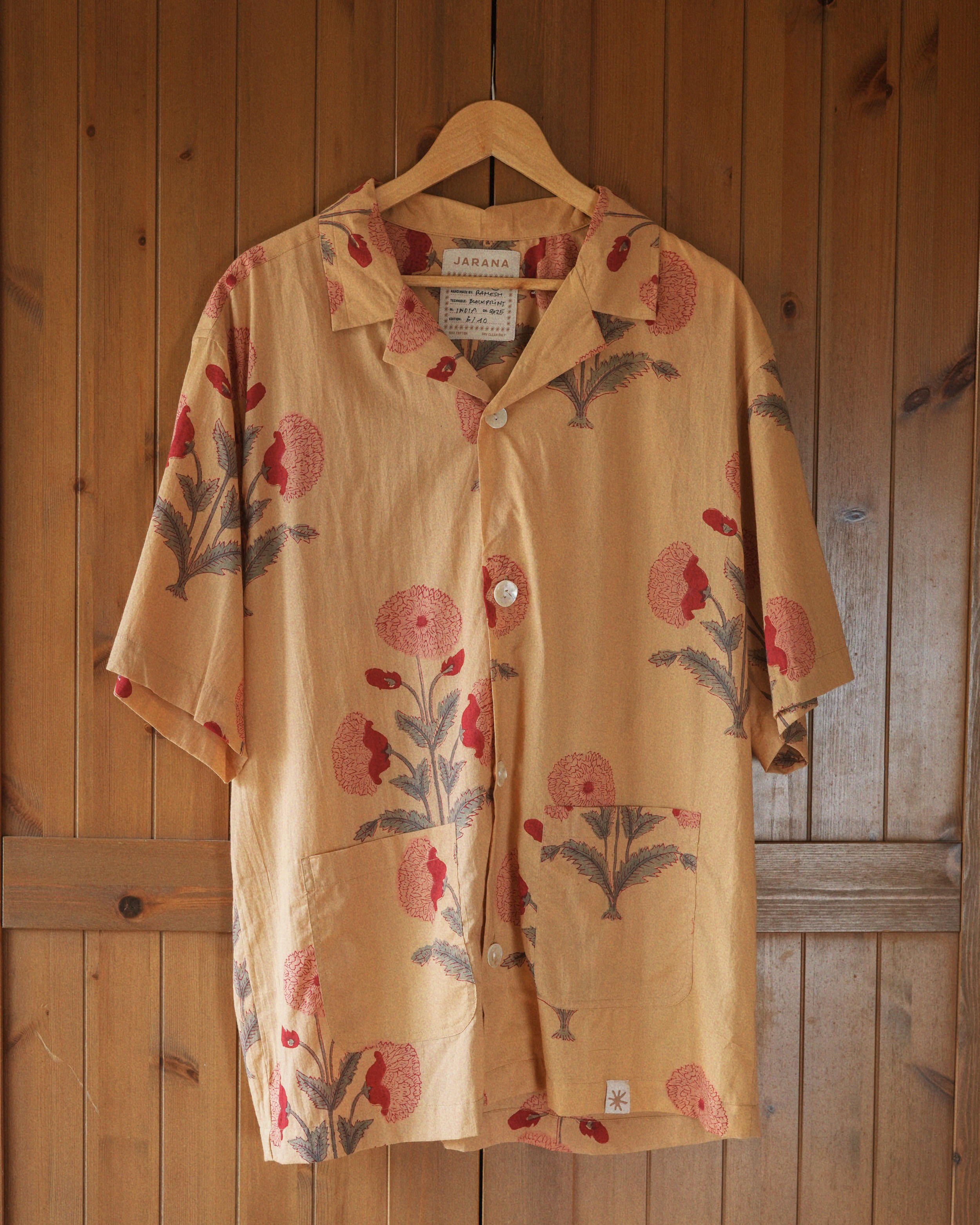 Short-sleeve yellow cotton shirt with red botanical block print, twin base pockets and shell buttons, hanging on wooden door.