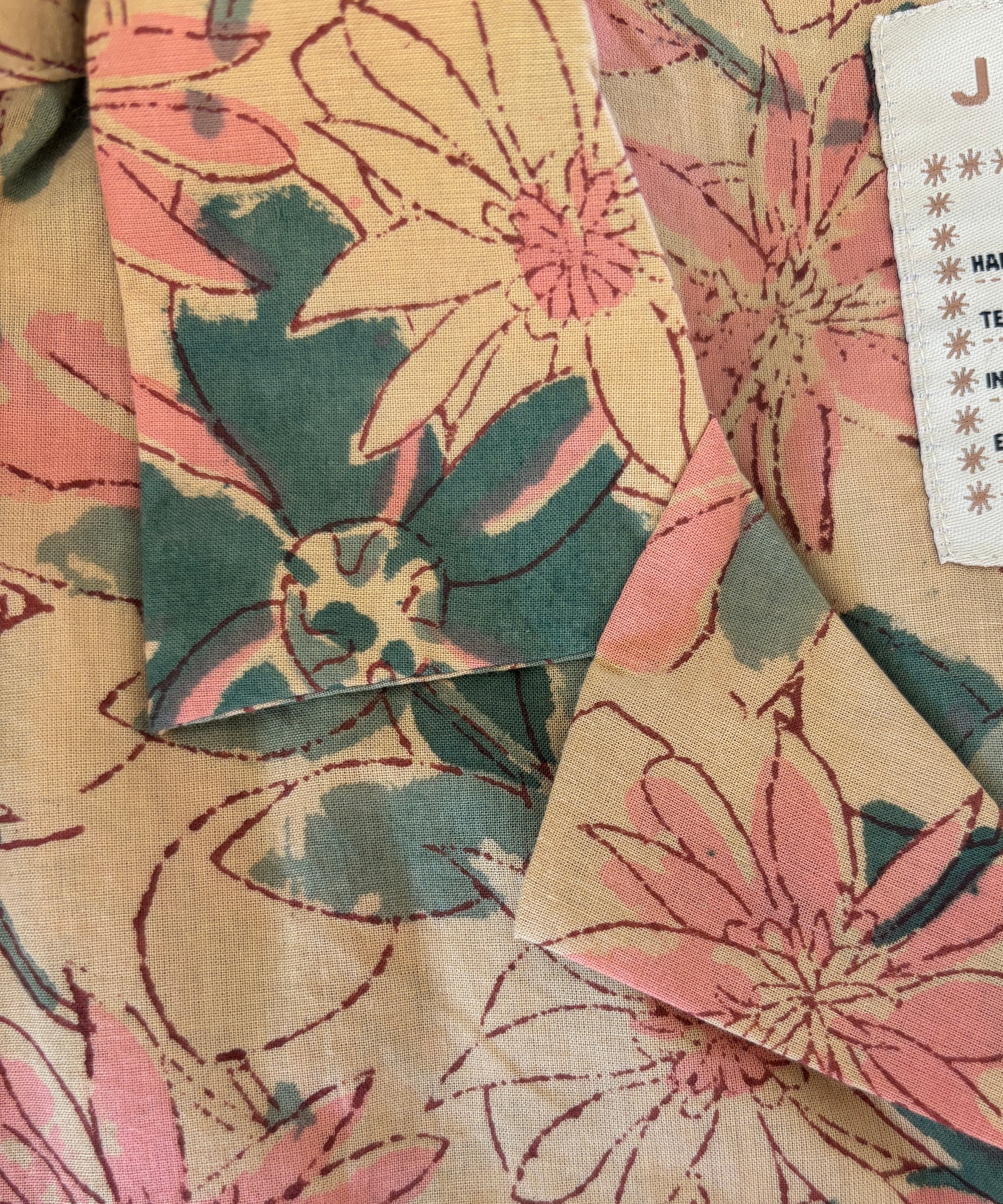 Close-up of Jarana shirt collar showing hand-sketched pink and green floral block print on beige cotton with visible size label.