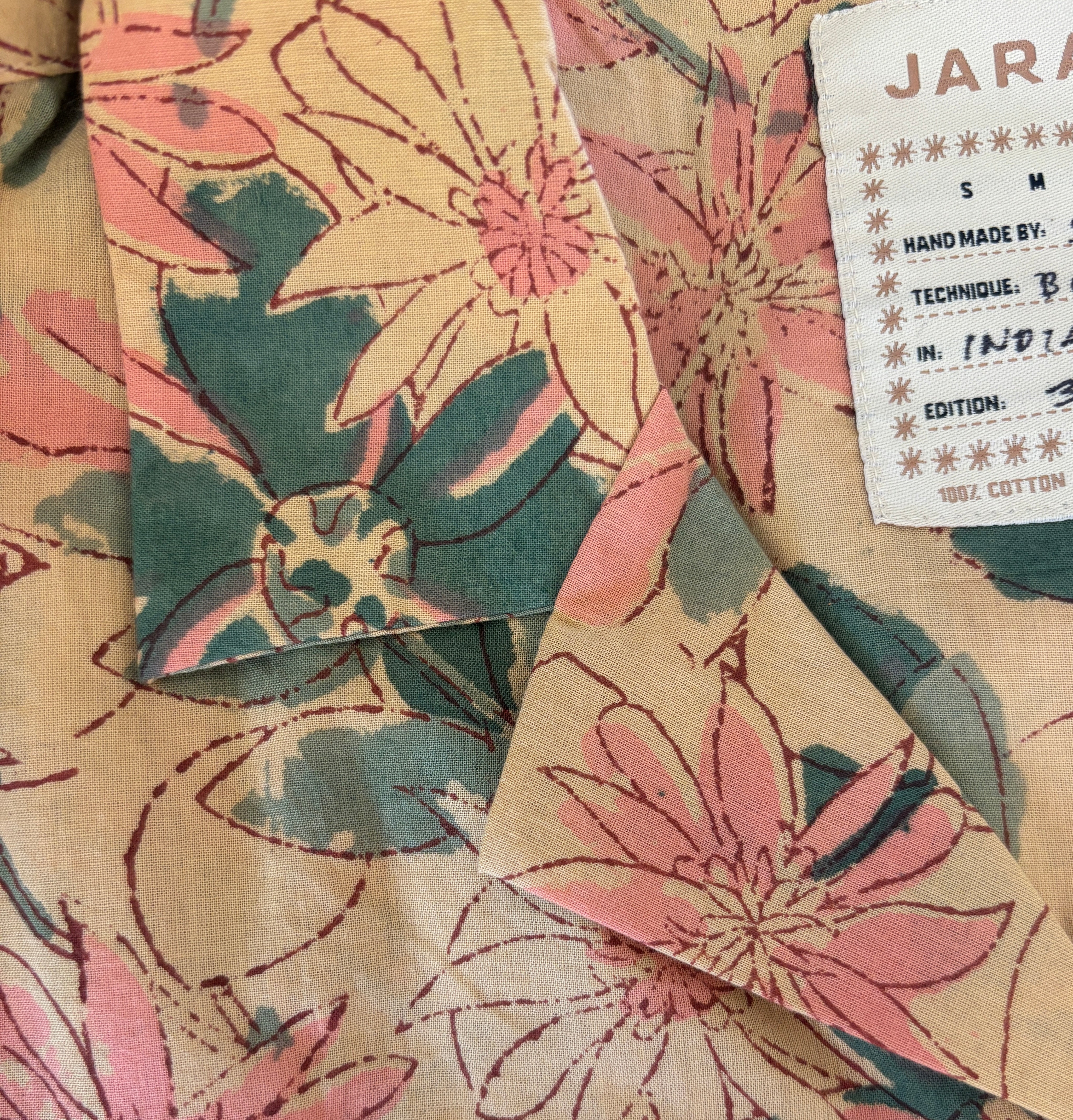 Close-up of Jarana shirt collar showing hand-sketched pink and green floral block print on beige cotton with visible size label.