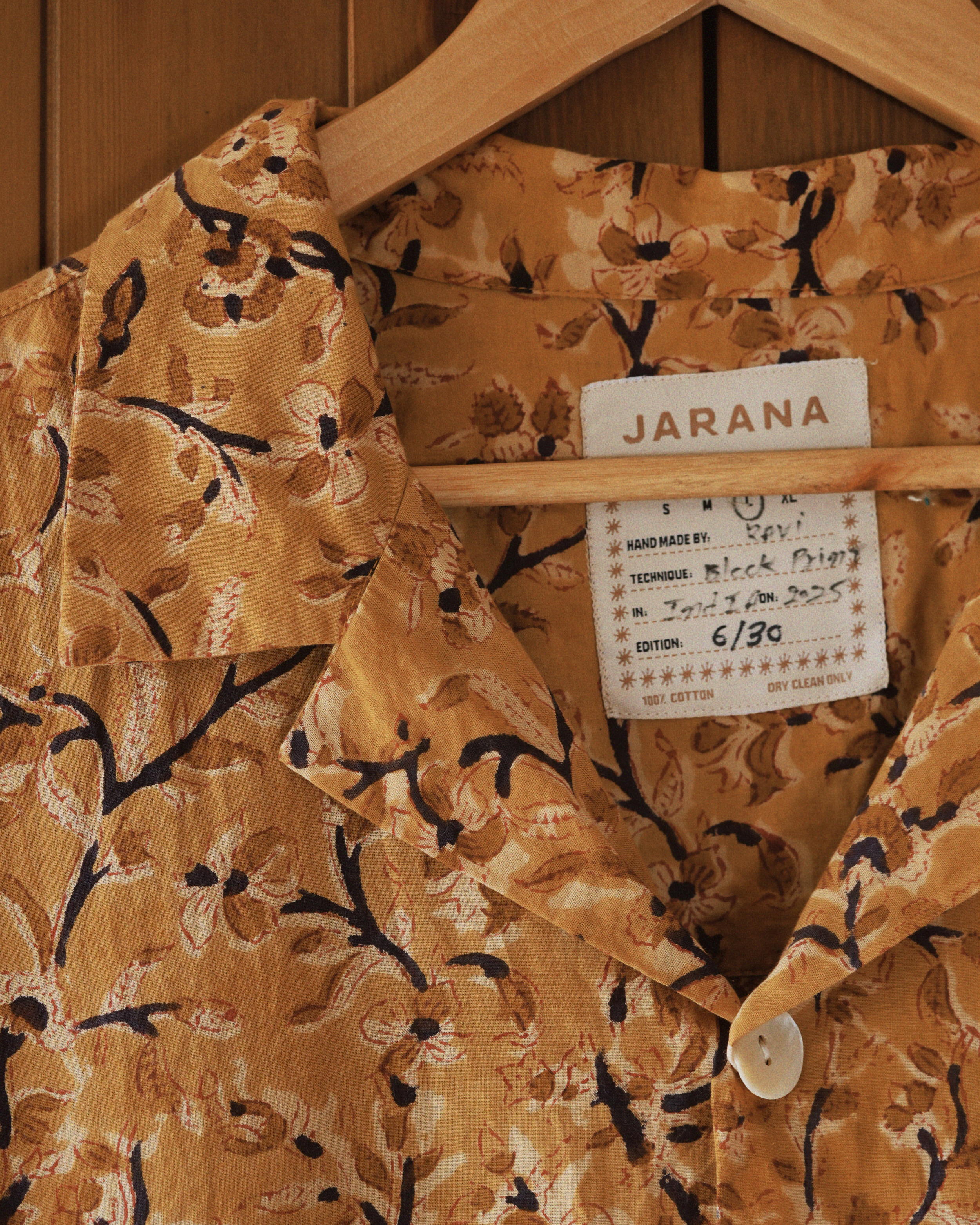Close-up of Ramaje short-sleeve shirt showing sepia floral block print, open resort collar, and Jarana label on wooden hanger.