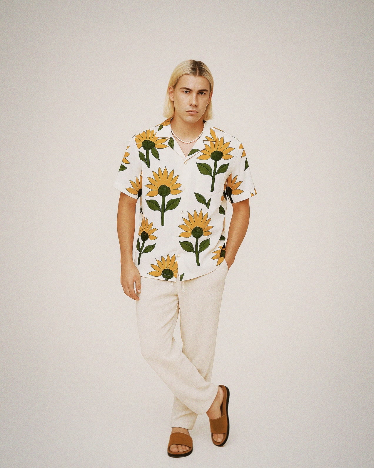 Full-body studio shot of a blond male model in a white sunflower-print cotton shirt, cream trousers, and sandals against a neutral backdrop.