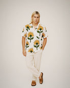 Full-body studio shot of a blond male model in a white sunflower-print cotton shirt, cream trousers, and sandals against a neutral backdrop.