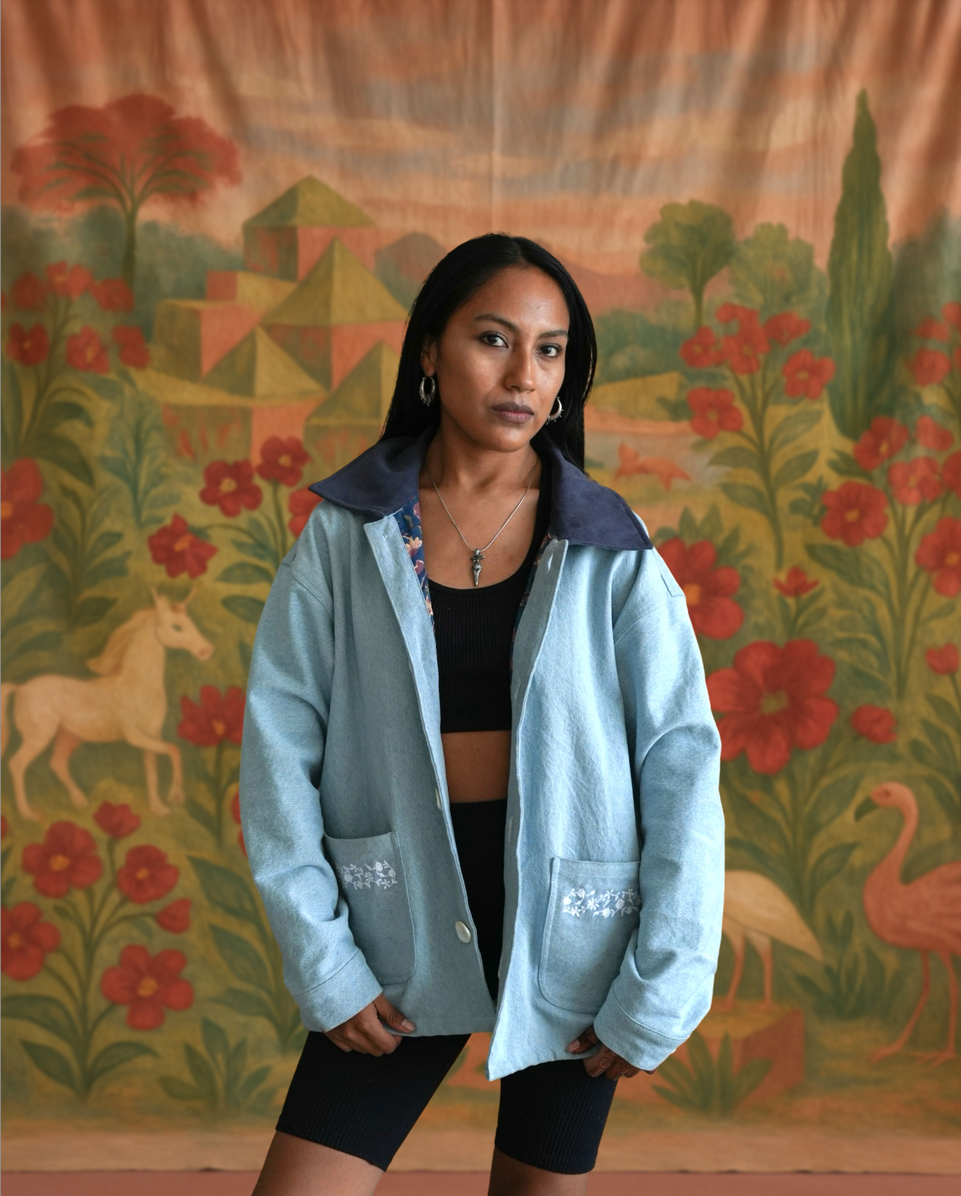 Model wearing sky blue denim jacket with dark corduroy collar and floral embroidered pockets by Jarana, styled against a backdrop of animals and red flowers.