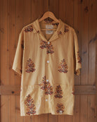 Full front view of the Flor Baja short-sleeve cotton shirt with twin base pockets and repeating red-gold floral motifs, hanging on a wooden hanger.