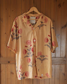 Short-sleeve yellow cotton shirt with red botanical block print, twin base pockets and shell buttons, hanging on wooden door.