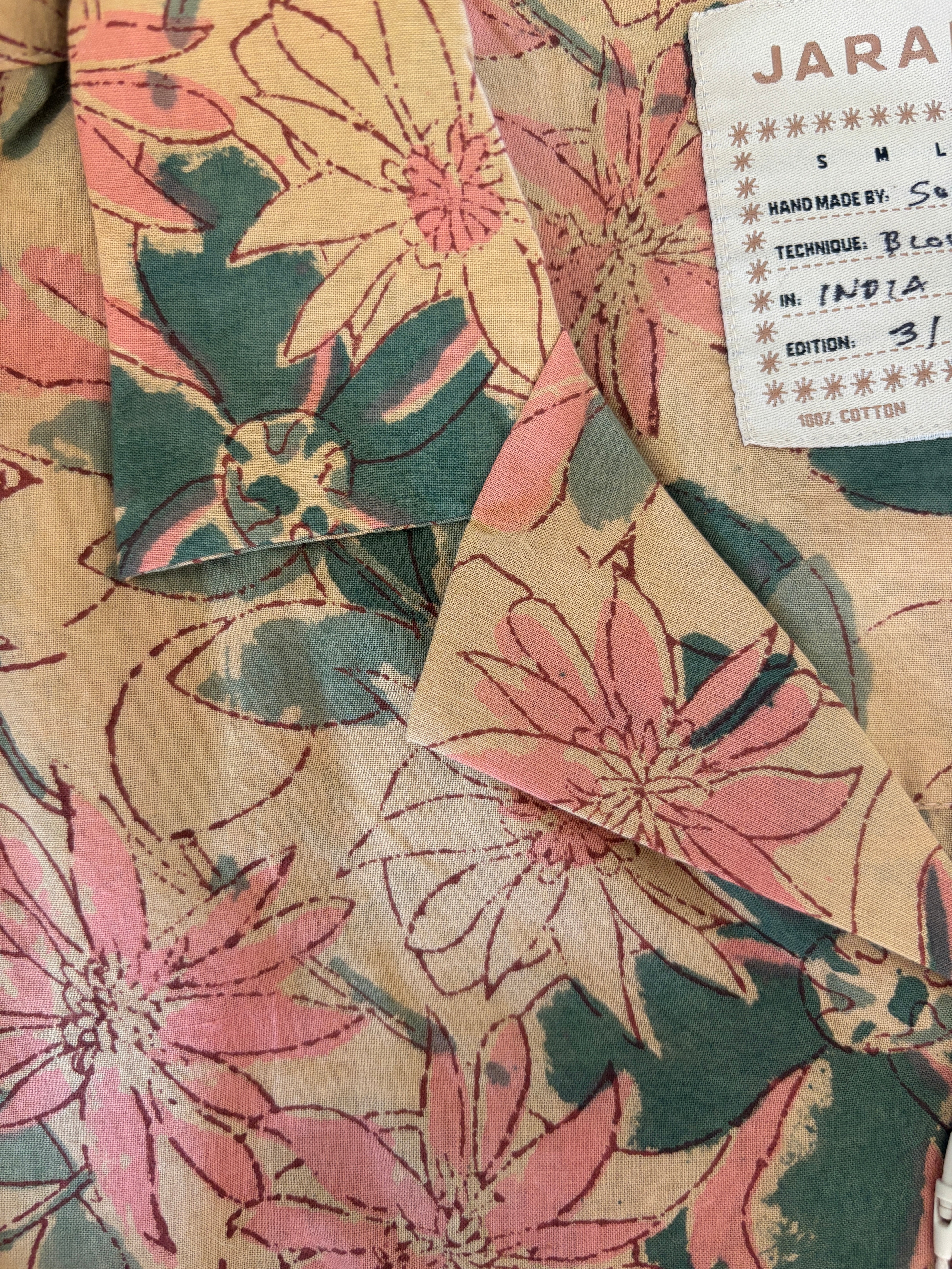 Close-up of Jarana shirt collar showing hand-sketched pink and green floral block print on beige cotton with visible size label.