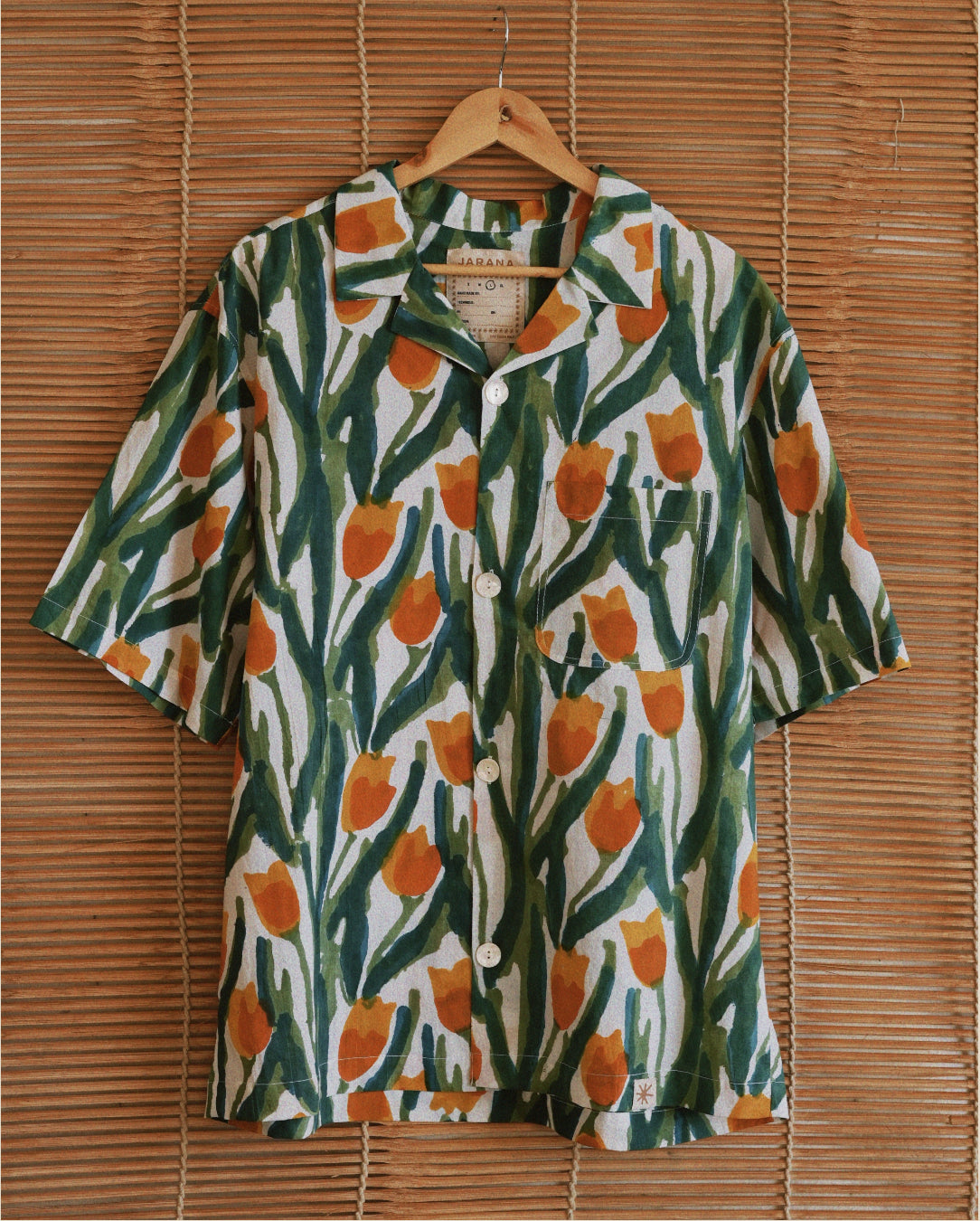 Full front view of Jarana tulip-print cotton shirt hanging on rattan backdrop, displaying orange flowers and leafy green pattern with white buttons.