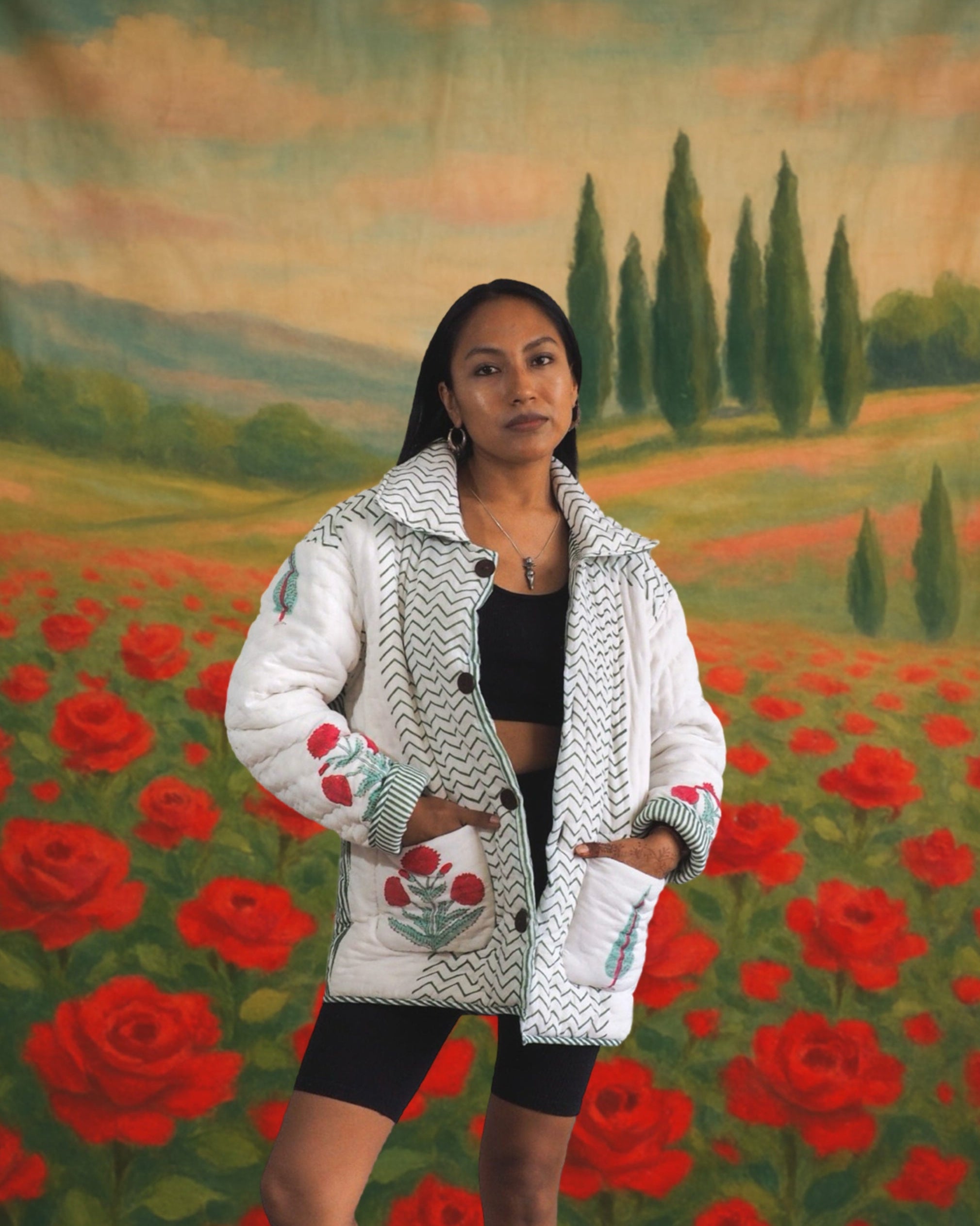 Model wearing white embroidered floral jacket by Jarana, standing in front of a romantic rose field backdrop with cypress trees