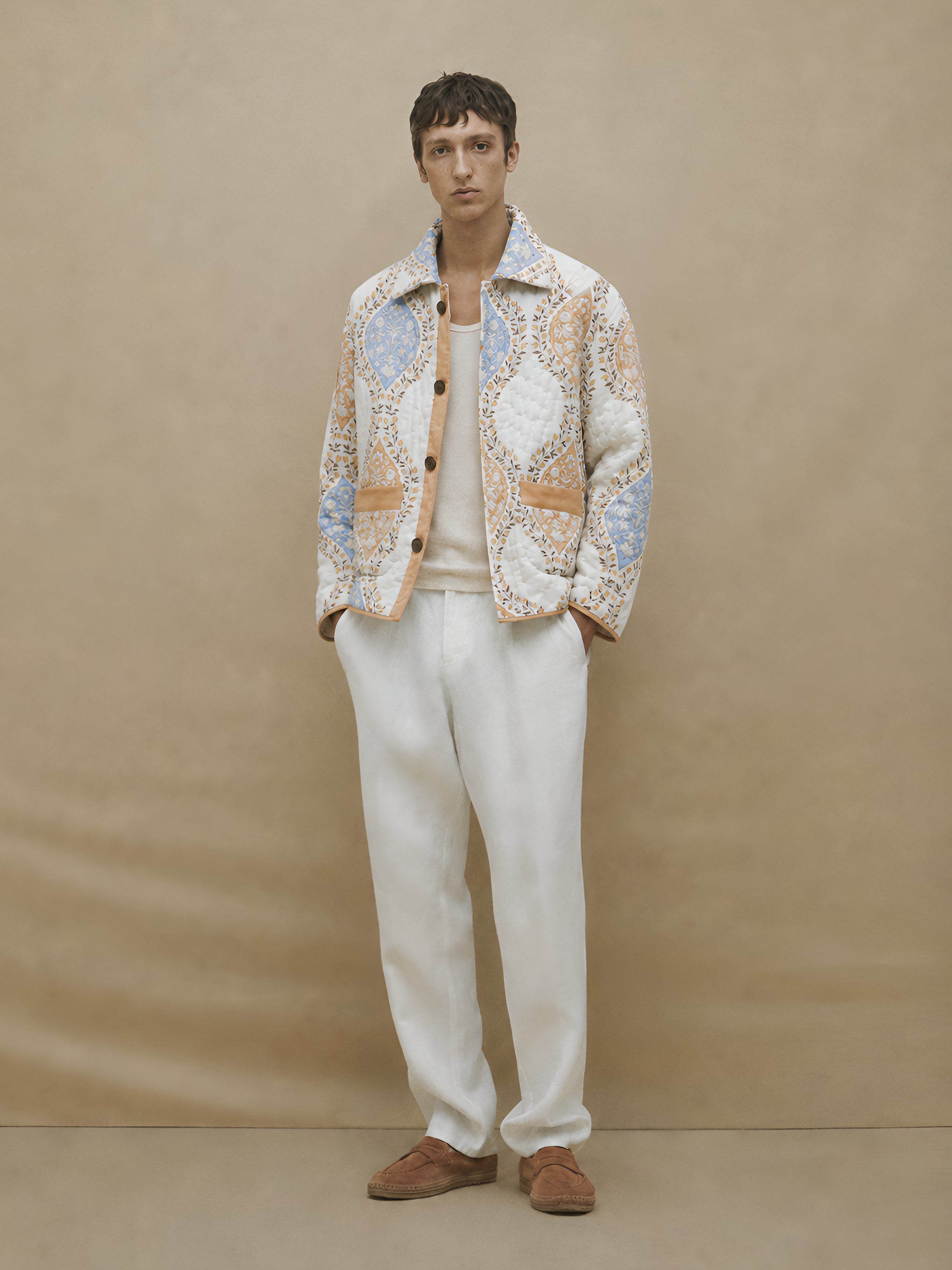 Man wearing a patterned jacket, white pants, and brown shoes against a beige background