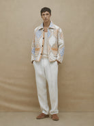Man wearing a patterned jacket, white pants, and brown shoes against a beige background