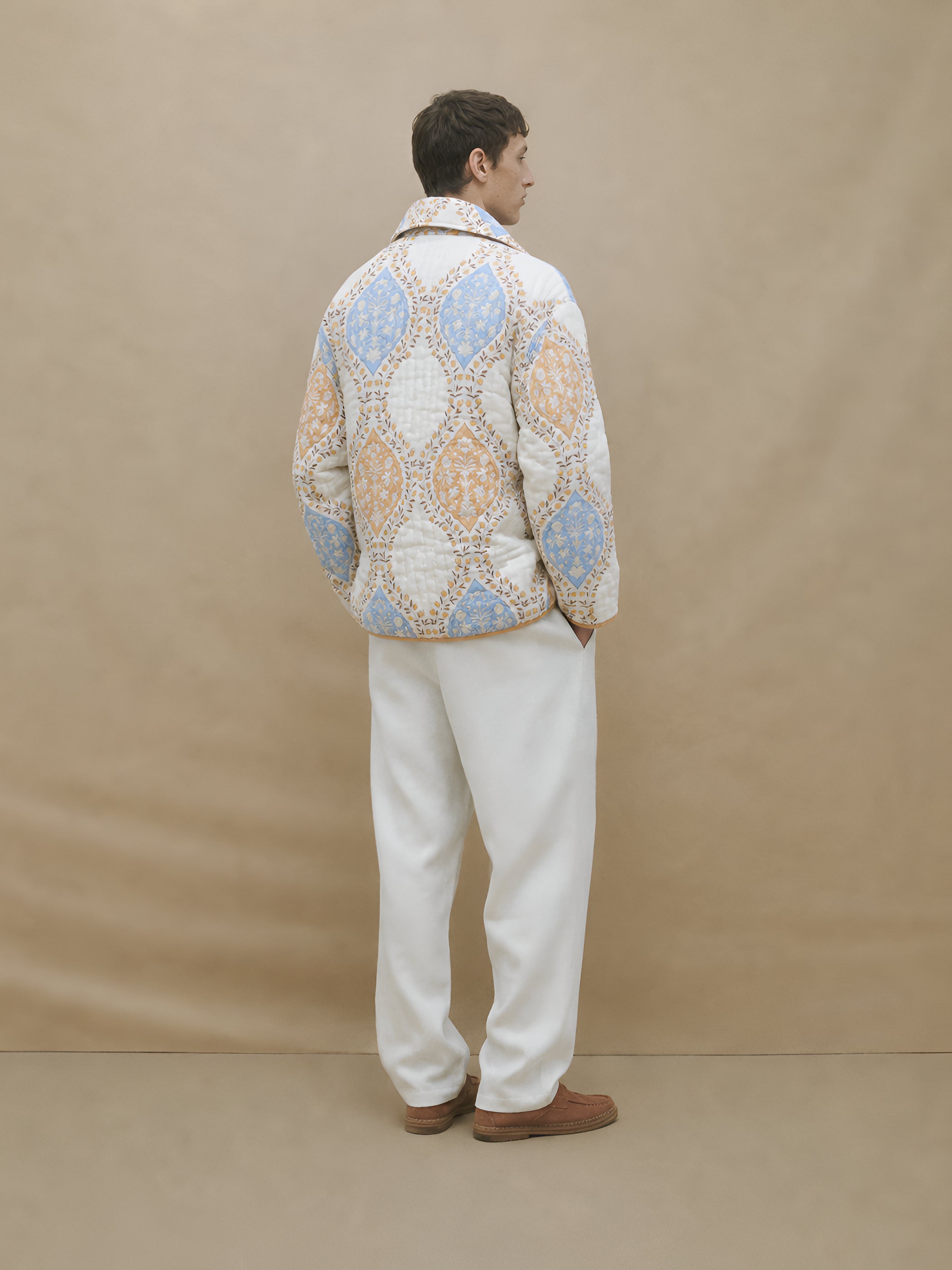 Person wearing a patterned jacket and white pants against a beige background
